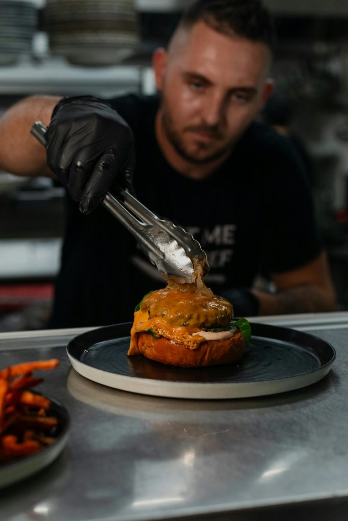 A chef skillfully prepares a gourmet cheeseburger with tongs in a professional kitchen setting.
