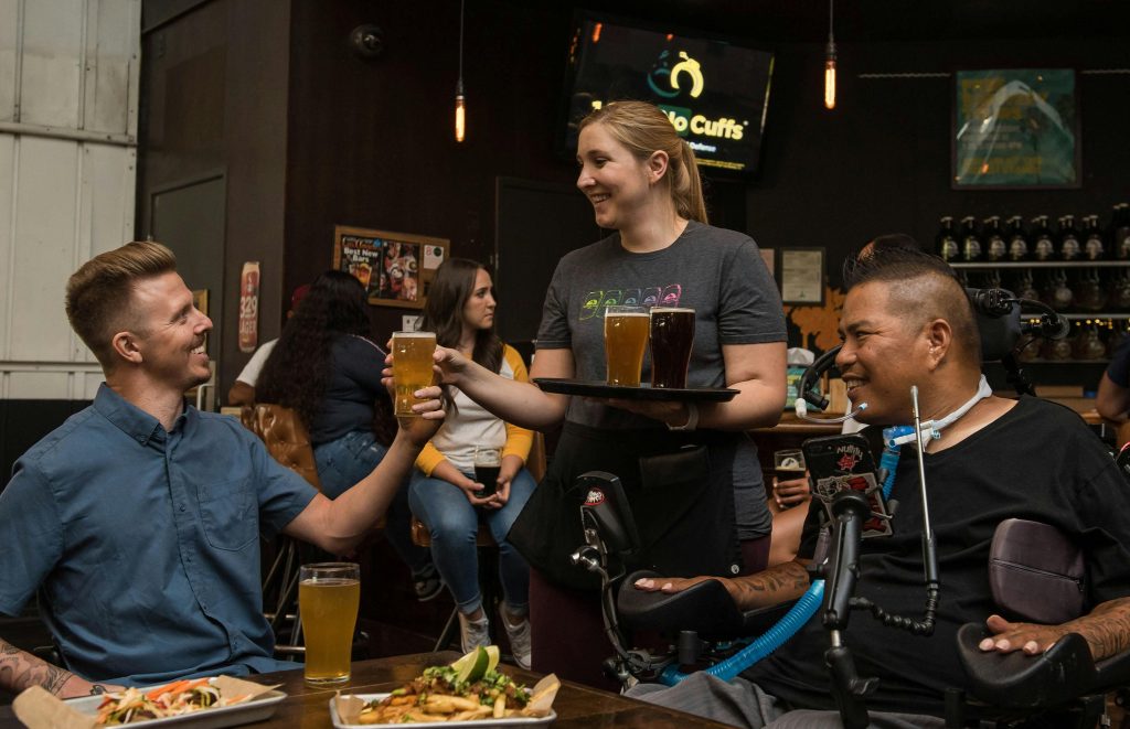 A waitress serves drinks to friends, including one in a wheelchair, in a lively bar setting.