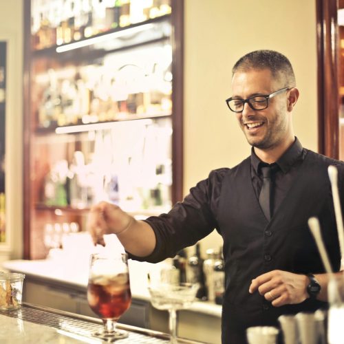 Happy young male barkeeper standing at counter and preparing alcohol cocktail for order while working in modern pub